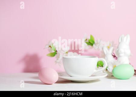 Tasse blanche avec thé ou café près rose et vert oeufs de Pâques, lapins et fleurs, sur une table blanche en béton, sur fond rose, avec des ombres dures. Banque D'Images