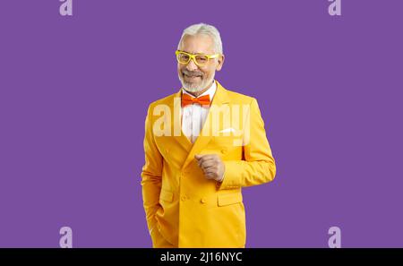 Homme senior heureux en costume jaune, noeud papillon et lunettes se tenant isolé sur fond violet Banque D'Images