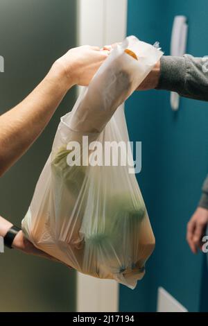 Gros plan vertical d'une petite cliente méconnue qui reçoit un sac d'épicerie du liveur devant le seuil de l'appartement. Banque D'Images