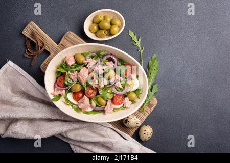 Vue de dessus de salade saine et savoureuse avec thon, arugula fraîche, tomates cerises, œuf de caille, oignon et olives vertes sur fond de béton noir Banque D'Images