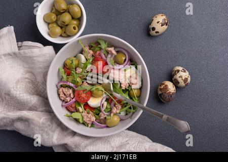 Vue de dessus de salade saine et savoureuse avec thon, arugula fraîche, tomates cerises, œuf de caille, oignon et olives vertes sur fond de béton noir Banque D'Images