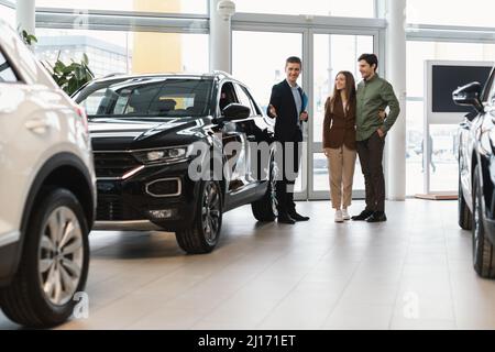 Vendeur confiant montrant une nouvelle voiture à un jeune couple caucasien chez un concessionnaire automobile Banque D'Images