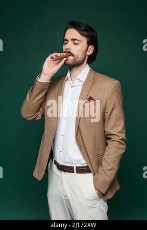 homme avec moustache fumant un cigare et debout avec la main dans la poche sur vert foncé Banque D'Images