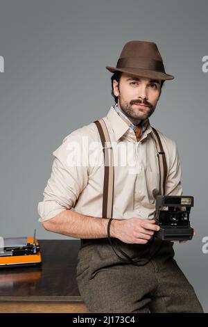 homme de style rétro vêtements et chapeau tenant appareil photo vintage et debout près du bureau avec machine à écrire isolée sur gris Banque D'Images