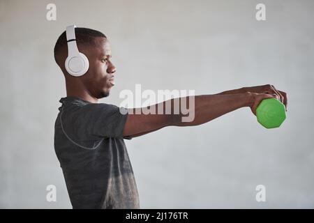 Vue latérale horizontale moyenne prise de vue d'un jeune Noir portant un casque blanc écoutant de la musique et faisant de l'exercice avec une haltère Banque D'Images