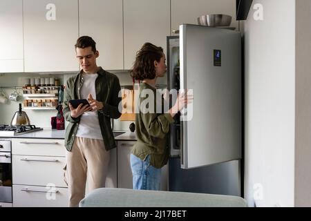 Jeune homme utilisant un PC tablette par une femme ouvrant le réfrigérateur dans la cuisine à la maison Banque D'Images