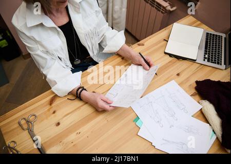Image rognée d'une femme d'affaires élégante et âgée coiffeuse assise sur une table en bois et tenant des croquis tout en travaillant dans son créateur Banque D'Images