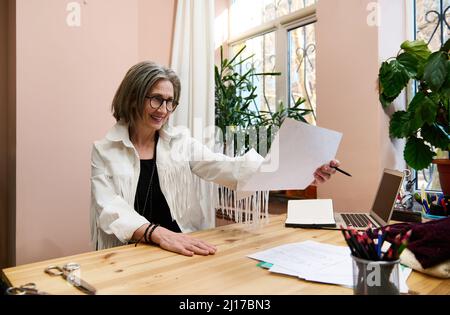 Femme d'affaires âgée et inspirée, la styliste féminine regarde des croquis dessinés tout en étant assise à une table dans son studio de design et en souriant avec che Banque D'Images