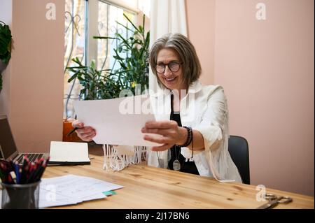 Femme d'affaires âgée inspirée femme designer de mode regarde des croquis dessinés pour des vêtements tout en étant assise à une table en bois dans son bureau de studio de design Banque D'Images