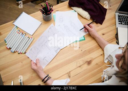 Vue de dessus. Gros plan d'une couturière de designer de mode âgée travaillant avec des croquis pour des vêtements assis sur un bureau en bois dans sa mode créative des Banque D'Images