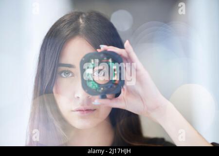 Jeune femme regardant à travers l'objectif ouvert de l'appareil photo au bureau Banque D'Images
