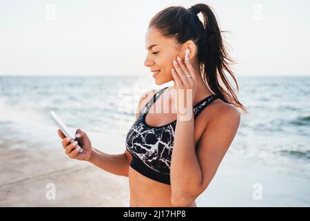 Femme souriante portant des écouteurs intra-auriculaires sans fil utilisant un smartphone à la plage Banque D'Images