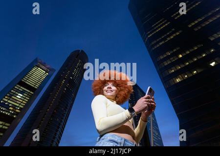 Femme souriante avec une coiffure afro à l'aide d'un smartphone devant des gratte-ciels Banque D'Images