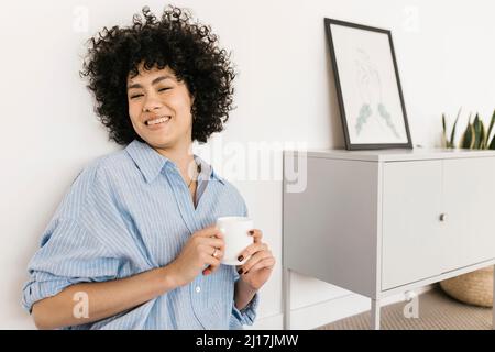 Une jeune femme heureuse avec une tasse de café assise près d'un mur blanc à la maison Banque D'Images