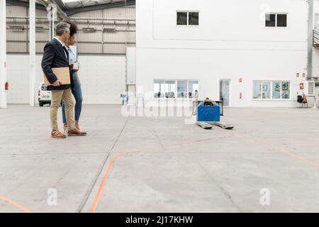 Homme d'affaires tenant un ordinateur portable debout avec une femme d'affaires regardant un transpalette en usine Banque D'Images