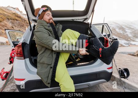 Jeune homme souriant dans le coffre de voiture mettant sur des bottes de ski Banque D'Images