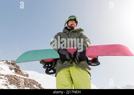 Jeune homme souriant debout avec snowboard par beau temps Banque D'Images