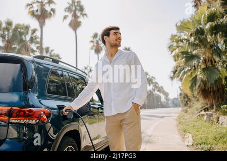 Homme souriant avec main dans la poche de recharge de voiture électrique au bord de la route Banque D'Images