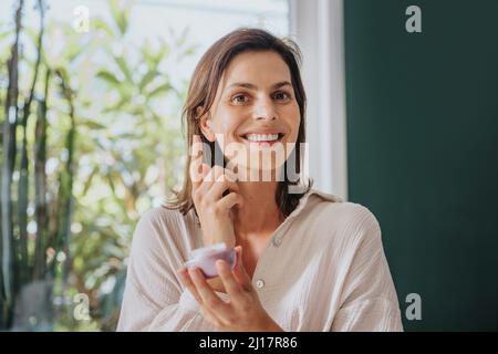 Femme souriante appliquant de la crème pour le visage à la maison Banque D'Images