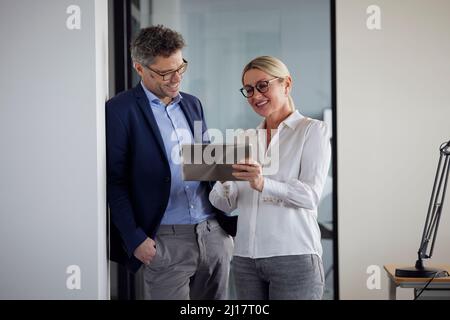 Un collègue explique au-dessus d'un Tablet PC à un homme d'affaires penché sur le mur avec la main dans la poche au bureau Banque D'Images