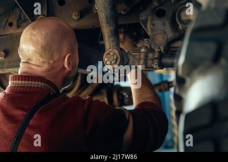 Le mécanicien ou mécanicien de voiture à chauve est assis sous la machinerie et répare la suspension du moteur de la voiture dans l'atelier. Entretien des camions lourds. Vue de l'arrière. Service de voiture. Arrière-plan. Banque D'Images