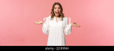 Surprise et heureuse jeune fille blonde réaliser qu'elle a gagné, recevoir vraiment de bonnes nouvelles, se répandre les mains sur le côté souriant et regarder étonné de l'appareil photo, debout rose Banque D'Images