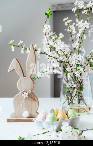 Lapin de Pâques en bois avec branche d'arbre en fleur avec fleurs dans le vase et œufs de pâques colorés dans le récipient sur la table blanche. Joyeuses Pâques Banque D'Images