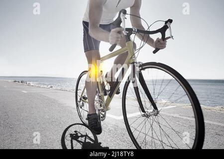 Traversez la douleur. Vue rognée d'un cycliste avec des problèmes de genou à vélo le long d'une route de l'océan. Banque D'Images