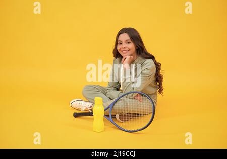 un enfant joyeux s'assoit dans des vêtements de sport avec raquette de badminton et bouteille d'eau sur fond jaune, détendez-vous Banque D'Images