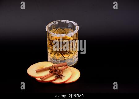 Un verre de cristal avec du cidre et du sucre autour des bords, à côté des pommes coupées, des bâtons de cannelle et de l'anis sur un fond noir. Gros plan. Banque D'Images