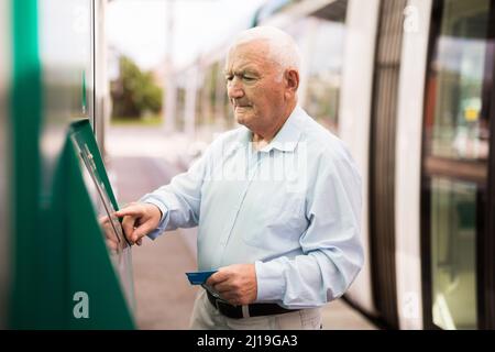 Homme senior utilisant un distributeur automatique de billets en se tenant à la station de tramway Banque D'Images