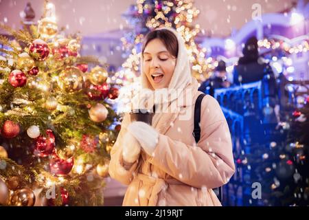 Bonne jeune femme avec une tasse de café chaud marche avec bokeh soirée lumière. Passer des vacances d'hiver sur la place Rouge, Moscou, Russie. Banque D'Images