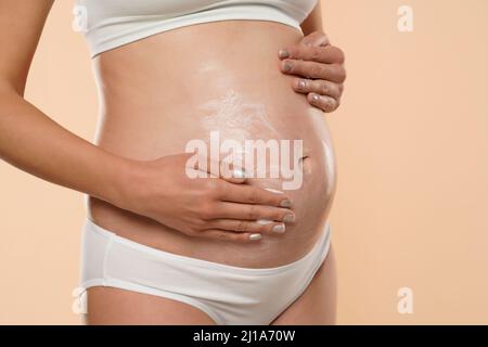 Vue rognée de la femme enceinte appliquant de la crème cosmétique sur son ventre pour éviter les marques d'étirement isolées sur beige Banque D'Images