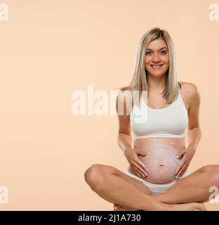 femme enceinte en position lotus appliquant de la crème cosmétique sur son ventre pour éviter les marques d'étirement sur fond beige Banque D'Images