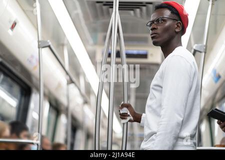 Portrait d'un étudiant noir bien pensé dans un chapeau rouge tenant une main courante avec une serviette en train de métro. Banque D'Images