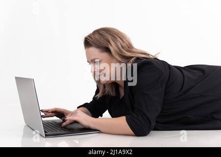 Portrait d'une belle femme d'âge moyen gras plus-taille portant un gilet noir, allongé sur un sol blanc, dactylographiant sur un ordinateur portable. Banque D'Images