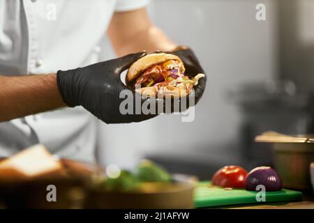 Vue rognée des mains du chef cuisinier dans des gants tenant un bon cheeseburger avec des légumes. Vue rapprochée de l'appétissant hamburger grillé dans le restaurant k Banque D'Images