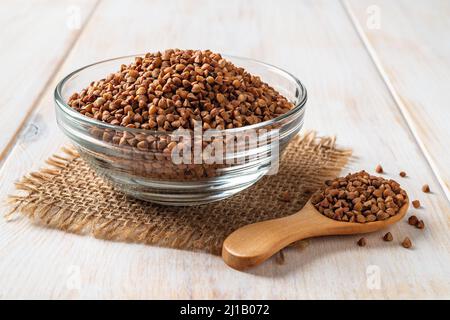Gros plan des gruaux de sarrasin cru dans un bol en verre et une cuillère en bois sur une table en bois blanc. Ingrédient pour la cuisson du porridge. Plats végétariens Banque D'Images