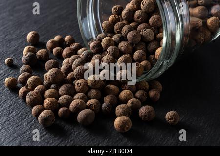 Des grains de poivre de jamaïque d'épices ont été déversés hors du pot de verre sur fond d'ardoise noire. Baies séchées de l'arbre Pimenta dioica. Épice biologique. Banque D'Images