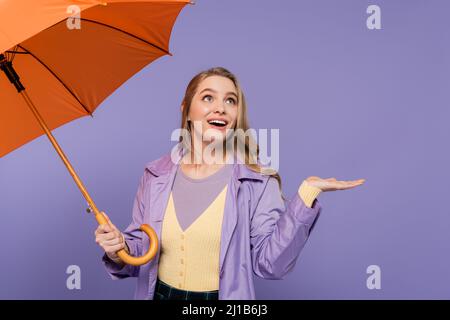 jeune femme gaie en trench pelage gestante tout en tenant un parapluie orange isolé sur le violet Banque D'Images