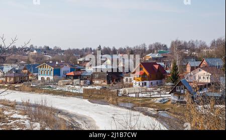 Une rivière incurvée, gelée s'étendant au loin, des maisons en bois sur la rive. Printemps, la neige fond, les flaques et l'herbe sèche tout autour. Jour, temps nuageux, lumière douce et chaude. Banque D'Images