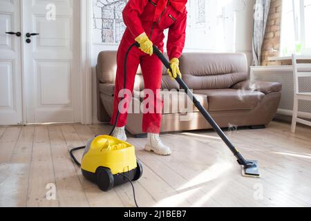 un produit de nettoyage en uniforme rouge nettoie le sol à l'aide de vapeur sortant de la brosse du balai. Banque D'Images