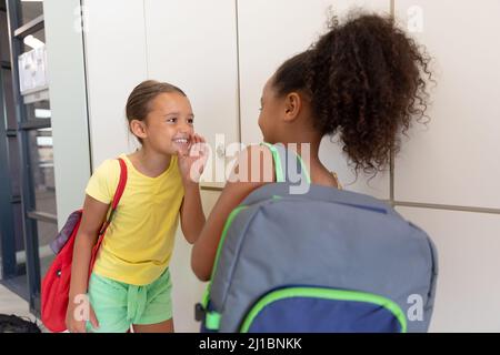 Des écolières élémentaires multiraciales commérages en persifant debout près de casiers dans le couloir de l'école Banque D'Images
