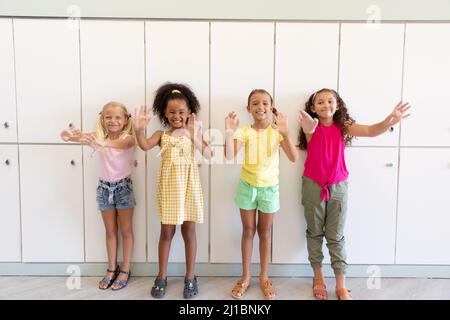 Portrait de filles d'école élémentaire multiraciale gaies gesturant tout en se tenant contre des casiers Banque D'Images