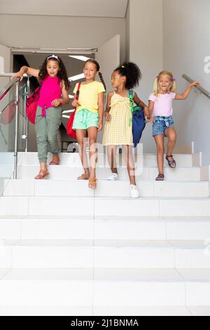 Filles d'école primaire multiraciale avec des sacs à dos sur les marches dans le bâtiment de l'école Banque D'Images