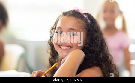 Portrait d'une jeune fille d'école élémentaire biraciale souriante assise avec la main sur le menton en classe Banque D'Images