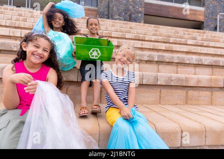 Portrait des filles d'école primaire multiraciale gaies avec des sacs à ordures assis sur les marches de l'école Banque D'Images