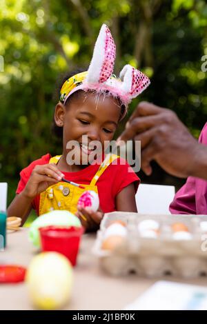 Souriant, fille et grand-père afro-américain dans des oreilles de lapin peignant des œufs dans la cour le jour de pâques Banque D'Images