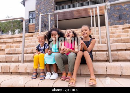 Les filles d'école primaire multiraciales et gaies sont assises sur des marches contre le bâtiment de l'école Banque D'Images