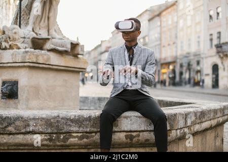 Homme afro-américain en costume d'affaires élégant assis dans la rue de la ville et à l'aide de lunettes VR. Concept de personnes, innovation et modes de vie modernes. Banque D'Images